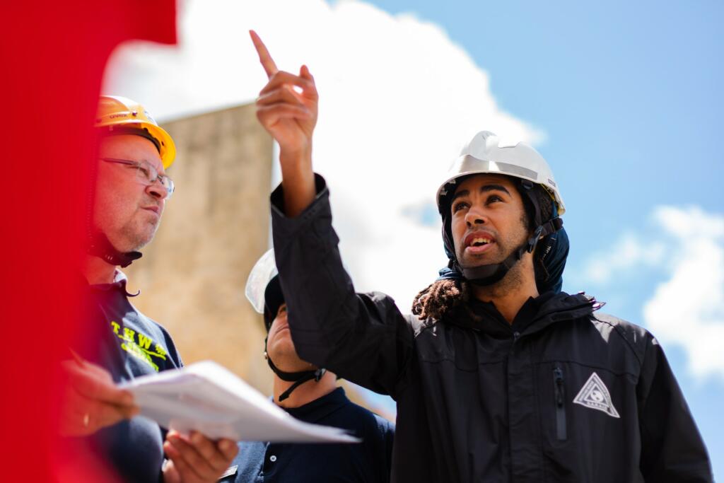 Man directing people at a job site, wearing a white hard hat and black jacket, pointing at something out of frame.